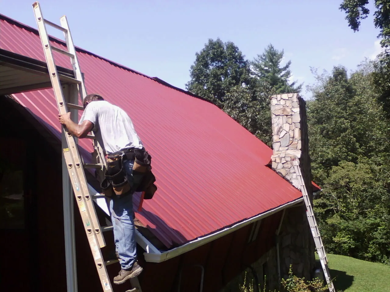 Roofer on a ladder beside a red metal roof and stone chimney — Soffit & Fascia Repair by Roofing Brothers in Warrington